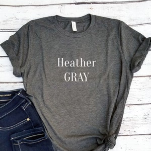May include: Heather Gray is printed in white text on a dark heather gray t-shirt. The t-shirt is folded and displayed on a white wooden surface.