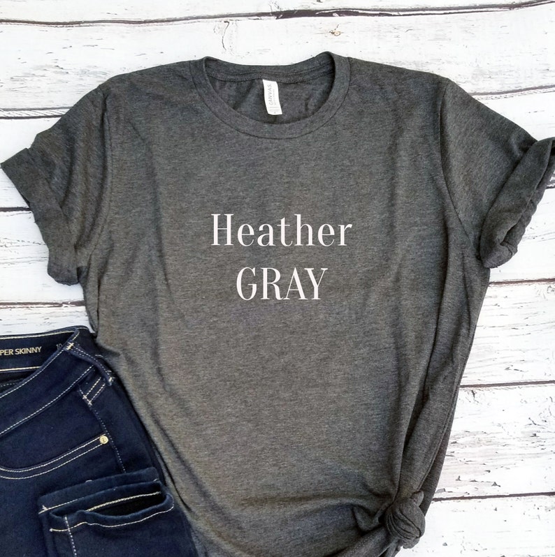 May include: Heather Gray is printed in white text on a dark heather gray t-shirt. The t-shirt is folded and displayed on a white wooden surface.