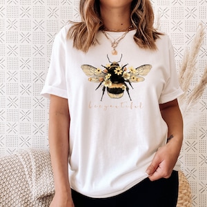 May include: White t-shirt with a graphic of a bee covered in yellow flowers. The text "bee-autiful" is printed below the bee.