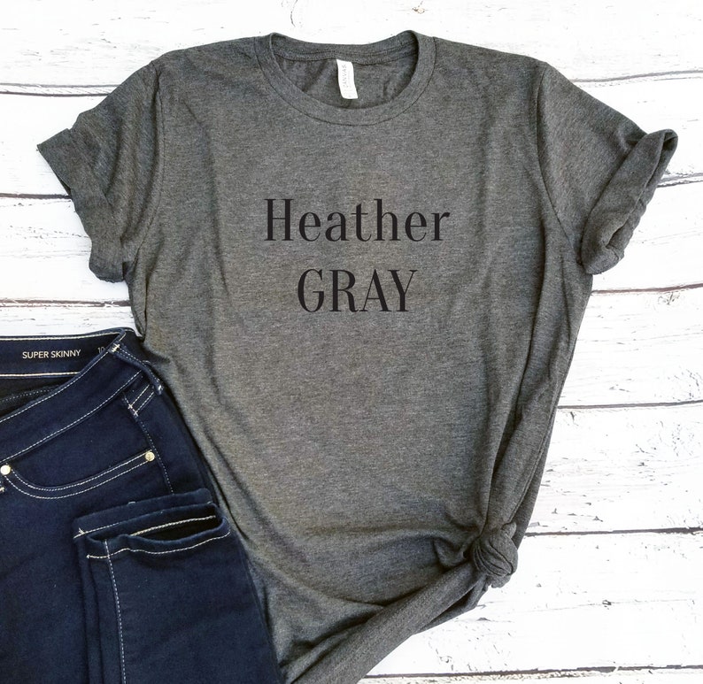 Puede incluir: Heather Gray est&aacute; impreso en negro sobre una camiseta gris jaspeada. La camiseta est&aacute; doblada y colocada sobre una superficie de madera blanca. Un par de pantalones vaqueros azules es parcialmente visible en la esquina inferior izquierda de la imagen.
