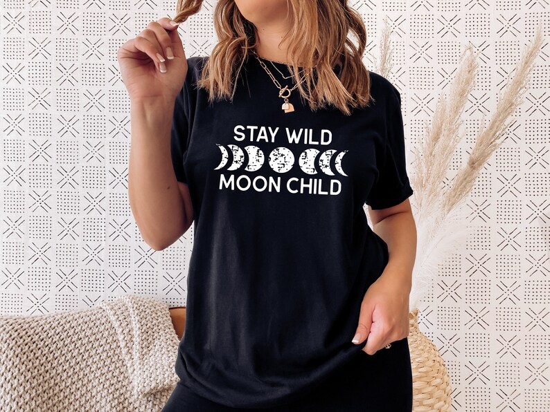 Peut inclure: T-shirt noir avec un texte blanc qui dit "Stay Wild Moon Child" et un graphique d'un croissant de lune dans diff&eacute;rentes phases.
