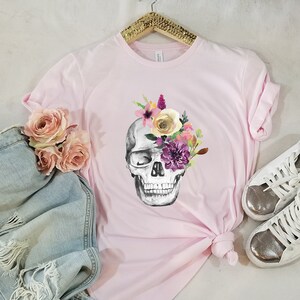 May include: A pink t-shirt with a floral skull graphic. The skull is gray and white with pink, purple, and yellow flowers.