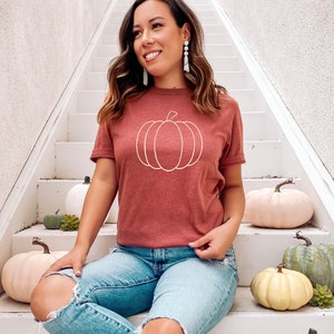 May include: A woman wearing a maroon t-shirt with a white outline of a pumpkin on the front. She is sitting on white stairs with several decorative pumpkins around her.