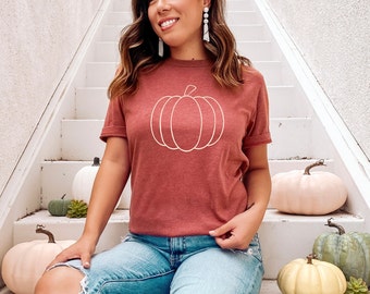 May include: A woman wearing a maroon t-shirt with a white outline of a pumpkin on the front. She is sitting on white stairs with several decorative pumpkins around her.