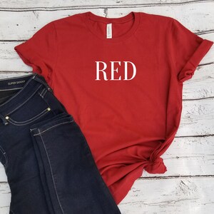 Puede incluir: Camiseta roja con la palabra "RED" impresa en blanco en la parte delantera. La camiseta se combina con unos vaqueros azul oscuro.
