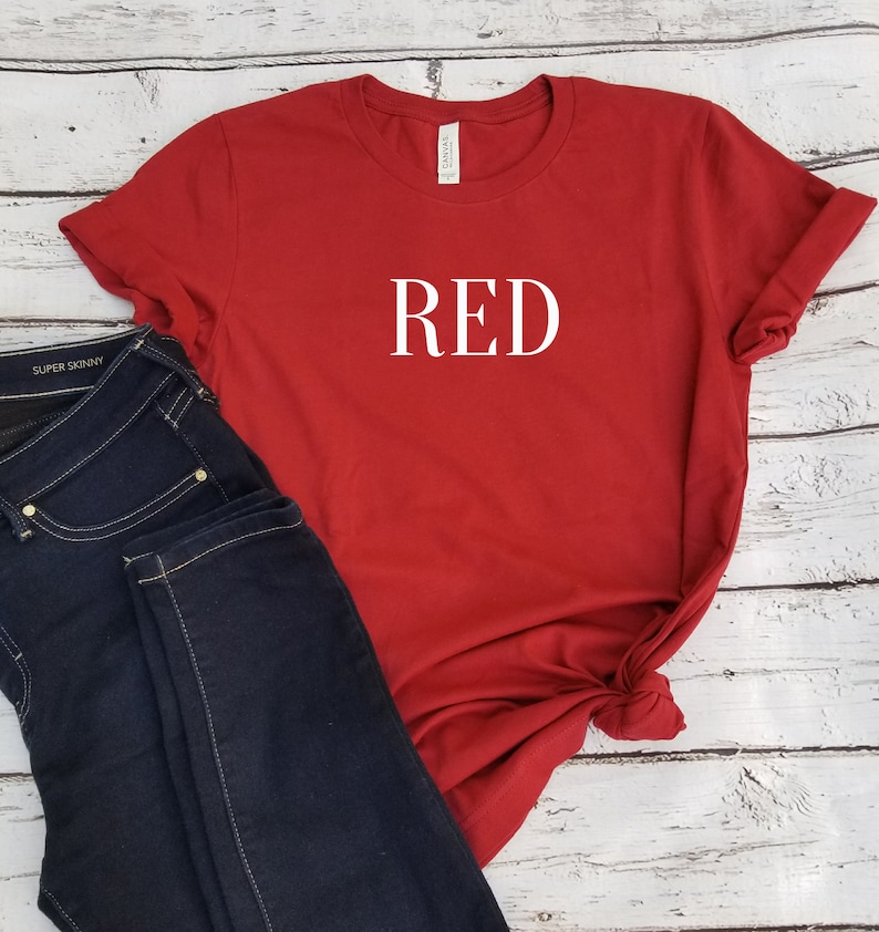 Puede incluir: Camiseta roja con la palabra "RED" impresa en blanco en la parte delantera. La camiseta se combina con unos vaqueros azul oscuro.