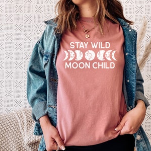 Peut inclure: Un t-shirt rose avec le texte "Stay Wild Moon Child" en lettres blanches. Le texte est accompagn&eacute; d'un graphique d'une lune dans diff&eacute;rentes phases.