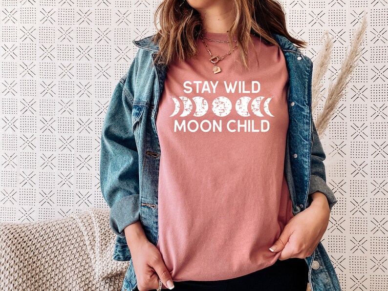 Peut inclure: Un t-shirt rose avec le texte "Stay Wild Moon Child" en lettres blanches. Le texte est accompagn&eacute; d'un graphique d'une lune dans diff&eacute;rentes phases.
