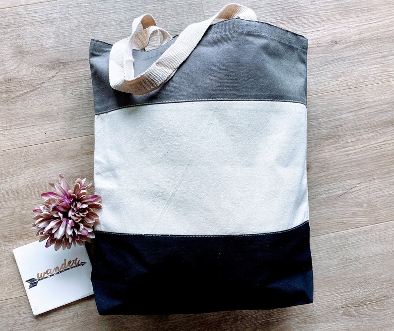 May include: A gray, white, and black striped canvas tote bag with two long handles. The bag is lying flat on a wooden surface.