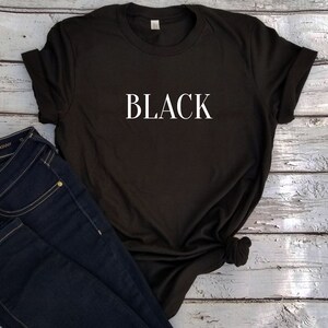 May include: Black t-shirt with the word "BLACK" printed in white on the front. The t-shirt is laying on a white wooden surface with a pair of blue jeans.