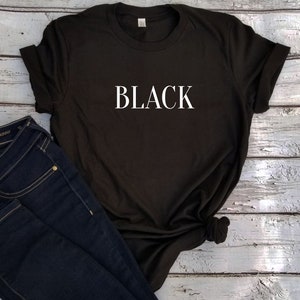 以下が含まれることがあります： 黒いTシャツに、白で「BLACK」とプリントされています。Tシャツは白い木製の表面に置かれ、青いジーンズが一緒にあります。