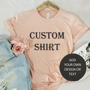 May include: A light pink t-shirt with the text "CUSTOM SHIRT" printed on the front. The text is in a black serif font. Below the text is a black circle with the text "ADD YOUR OWN DESIGN OR TEXT" in white.