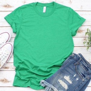 May include: A plain green t-shirt with rolled up sleeves. The t-shirt is laying on a white wooden surface with a pair of distressed denim shorts and white sneakers.