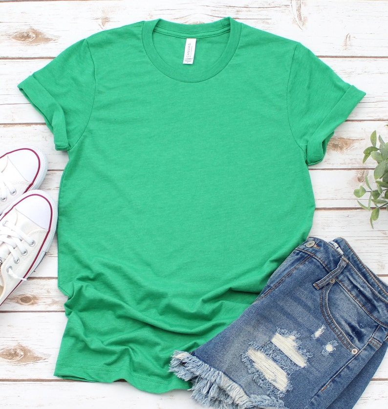 May include: A plain green t-shirt with rolled up sleeves. The t-shirt is laying on a white wooden surface with a pair of distressed denim shorts and white sneakers.