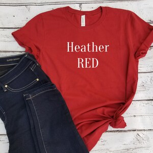 May include: Red t-shirt with the text "Heather RED" printed in white on the front. The shirt is paired with dark blue jeans.