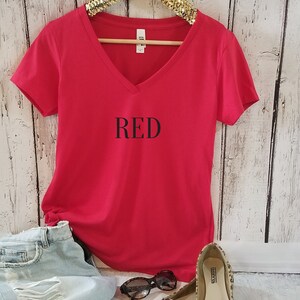 May include: Red V-neck t-shirt with the word "RED" printed in black on the front. The shirt is displayed on a hanger with a pair of leopard print flats and sunglasses.