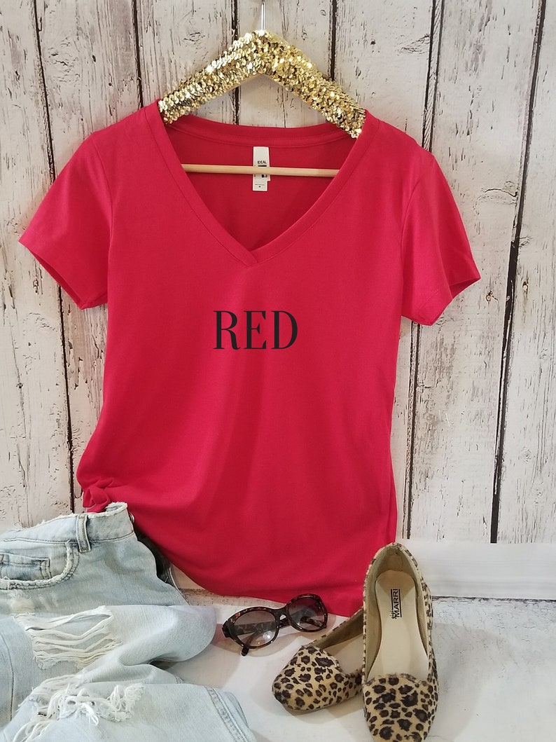 May include: Red V-neck t-shirt with the word "RED" printed in black on the front. The shirt is displayed on a hanger with a pair of leopard print flats and sunglasses.