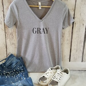 May include: Gray V-neck t-shirt with the word "GRAY" printed on the front. The shirt is paired with blue jeans, white sneakers, and sunglasses.