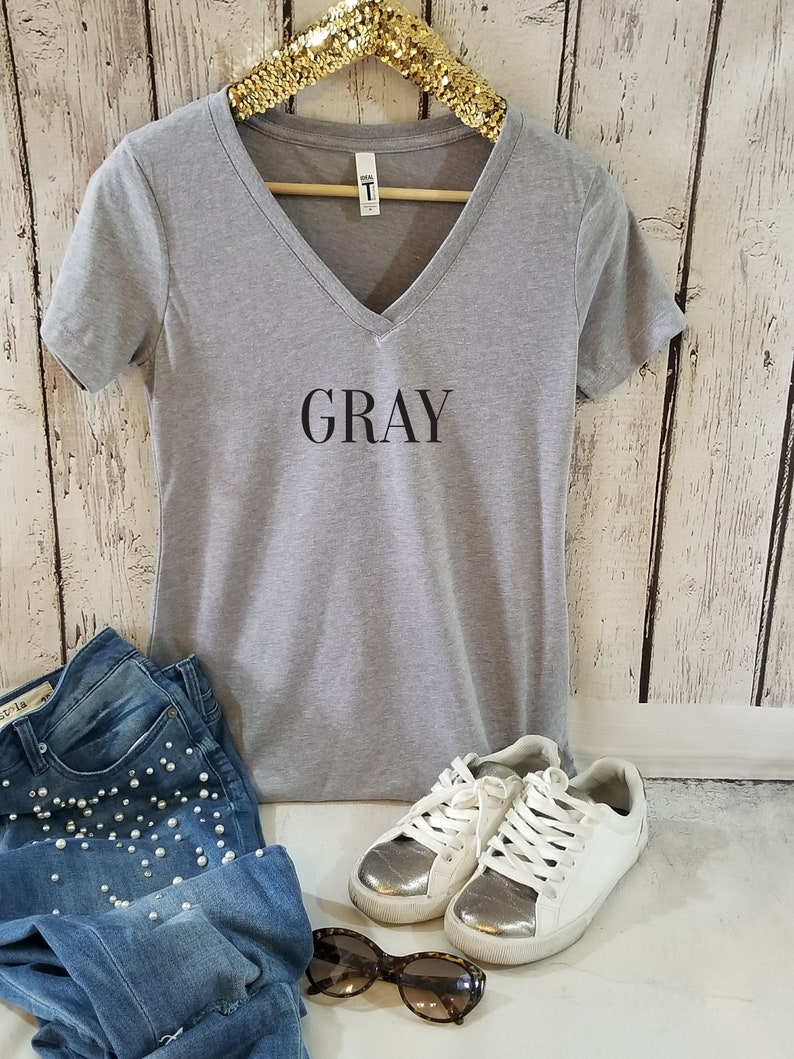May include: Gray V-neck t-shirt with the word "GRAY" printed on the front. The shirt is paired with blue jeans, white sneakers, and sunglasses.