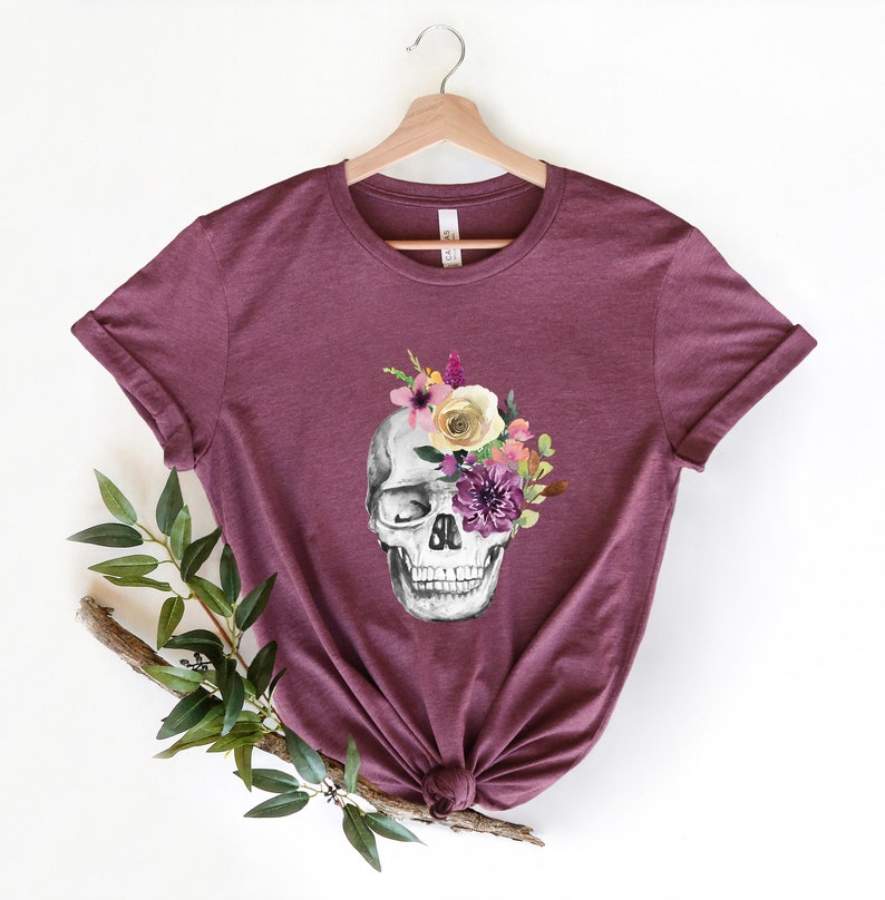 May include: A maroon t-shirt with a white skull design. The skull has a floral arrangement covering one eye socket. The flowers are purple, pink, yellow, and white.