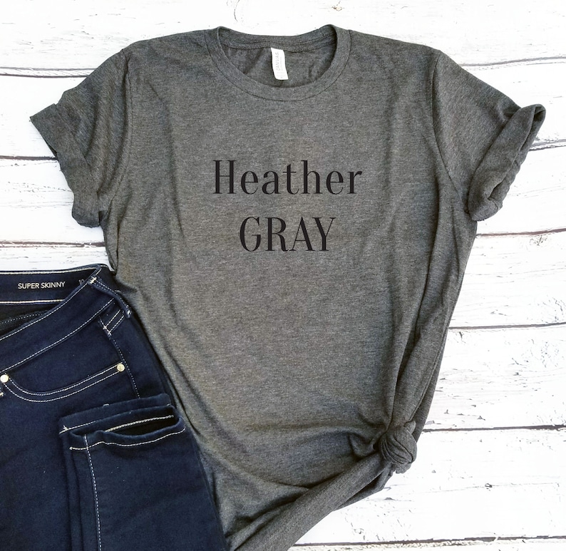 May include: Heather Gray is printed in black on a heather gray t-shirt. The t-shirt is folded and laid on a white wooden surface. A pair of blue jeans is partially visible in the lower left corner of the image.