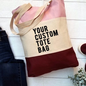 May include: A pink, beige, and burgundy canvas tote bag with the text "YOUR CUSTOM TOTE BAG" printed in black.