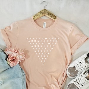 May include: A pink t-shirt with the word "ABRACADABRA" spelled out in white letters in a grid pattern.