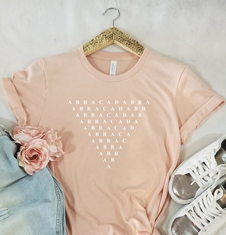 May include: A pink t-shirt with the word "ABRACADABRA" spelled out in white letters in a grid pattern.