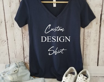 Op de afbeelding: Een marineblauw v-hals T-shirt met de tekst "Custom DESIGN Shirt" in witte letters. Het shirt hangt aan een gouden hanger en wordt getoond op een witte houten achtergrond.