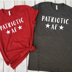 May include: Two t-shirts with the text "PATRIOTIC AF" and stars printed on them. The shirt on the left is red and the shirt on the right is gray.