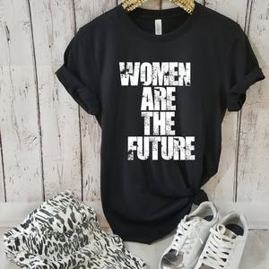 May include: Black t-shirt with white text that reads "Women Are The Future". The shirt is on a hanger and is styled with a leopard print skirt, white sneakers, and sunglasses.