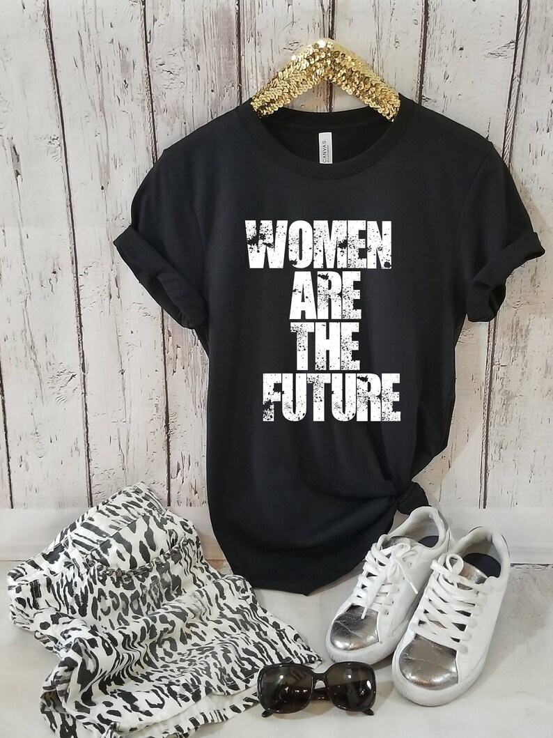 May include: Black t-shirt with white text that reads "Women Are The Future". The shirt is on a hanger and is styled with a leopard print skirt, white sneakers, and sunglasses.