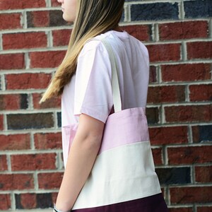 May include: A person wearing a pink and white striped t-shirt and blue jeans is holding a pink, white, and burgundy striped tote bag.