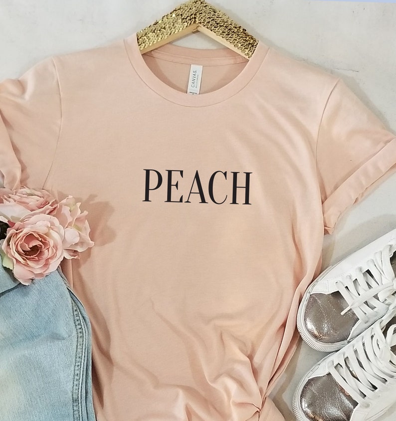 以下が含まれることがあります： ピーチ色のTシャツに、黒い文字で「PEACH」とプリントされています。Tシャツは白い表面に置かれ、銀色のアクセントが付いた白いスニーカーが置かれています。
