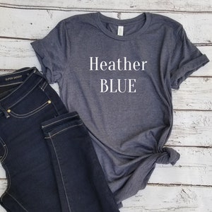 May include: Heather blue text printed on a dark gray t-shirt. The t-shirt is laying on a white wooden surface with a pair of dark blue jeans.