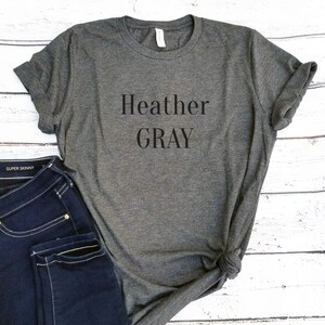 May include: Heather Gray is printed in black on a heather gray t-shirt. The t-shirt is folded and laid on a white wooden surface. A pair of blue jeans is partially visible in the bottom left corner of the image.