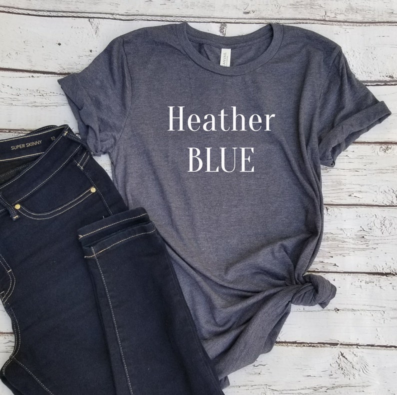May include: Heather blue text printed on a dark gray t-shirt. The t-shirt is laying on a white wooden surface with a pair of dark blue jeans.