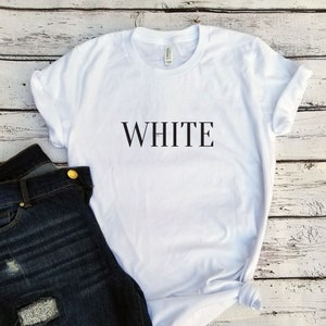 May include: White t-shirt with the word "WHITE" printed in black on the front. The shirt is laying on a white wooden surface with a pair of blue jeans to the left.
