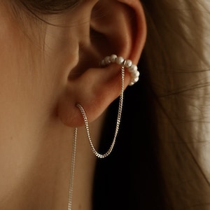 Może przedstawiać: Delikatny zestaw nausznicy i kolczyków. Nausznica zdobiona jest małymi białymi perłami, a kolczyk ma długi, zwisający srebrny łańcuszek. Biżuteria jest stylowym dodatkiem.