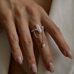 Puede incluir: Un anillo de plata con una perla blanca grande y un dije de perla colgante. El anillo está en un dedo con esmalte de uñas rosa claro y detalles dorados.