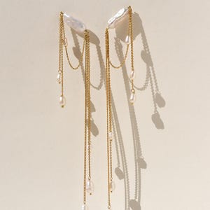 Può includere: Un paio di orecchini eleganti con grandi perle bianche dalla forma irregolare sospese a delicate catene d'oro. Perle più piccole sono intervallate lungo le catene, creando un effetto a cascata. Sfondo neutro.