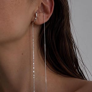 May include: A silver ear cuff featuring a wavy design connected to a long, delicate chain that hangs down. A second, thin, straight silver chain also drapes from the ear, creating a layered, dangling effect that extends past the jawline.