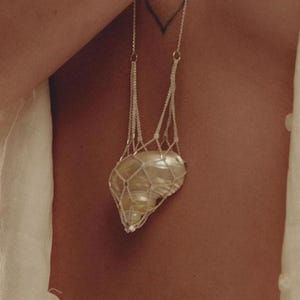 Puede incluir: Un collar plateado con un colgante que contiene una piedra grande y clara en una red fina. El collar se lleva sobre la piel, con un tatuaje de corazón visible. Se lleva una parte superior blanca. Joyería única.