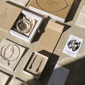 Pu&ograve; includere: Un assortimento di gioielli, tra cui collane, bracciali e orecchini, &egrave; esposto in scatole e sacchetti aperti. I gioielli sono presentati su una superficie chiara, con il marchio "ANNA KOVA jewelry" visibile sulle scatole e sulle carte.