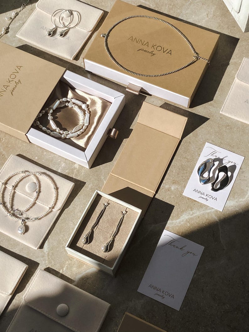 Pu&ograve; includere: Un assortimento di gioielli, tra cui collane, bracciali e orecchini, &egrave; esposto in scatole e sacchetti aperti. I gioielli sono presentati su una superficie chiara, con il marchio "ANNA KOVA jewelry" visibile sulle scatole e sulle carte.