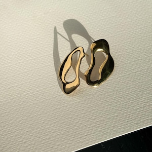K&ouml;nnte beinhalten: Ein Paar goldfarbene, abstrakt geformte Ohrringe. Die Ohrringe haben ein flie&szlig;endes, organisches Design mit unregelm&auml;&szlig;igen Kanten und einer polierten Oberfl&auml;che. Die Ohrringe werden auf einer wei&szlig;en Oberfl&auml;che mit einem Schatten an der Seite pr&auml;sentiert.