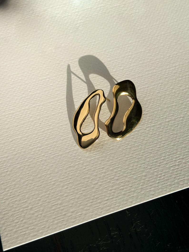 K&ouml;nnte beinhalten: Ein Paar goldfarbene, abstrakt geformte Ohrringe. Die Ohrringe haben ein flie&szlig;endes, organisches Design mit unregelm&auml;&szlig;igen Kanten und einer polierten Oberfl&auml;che. Die Ohrringe werden auf einer wei&szlig;en Oberfl&auml;che mit einem Schatten an der Seite pr&auml;sentiert.