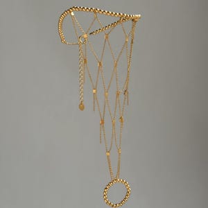 Könnte beinhalten: Eine goldfarbene Handkette mit einem perlenbesetzten Ring und einem filigranen Kettennetz-Design. Die Kette hat kleine quadratische Akzente und eine perlenbesetzte Manschette. Der Schmuck hängt vor einem grauen Hintergrund.