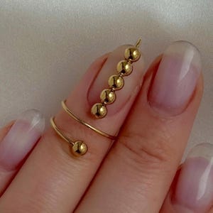 Peut inclure: Une bague dorée avec un design en spirale et une série de petites perles rondes. La bague s'enroule autour du doigt, les perles étant alignées sur le dessus. Un bijou délicat.