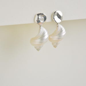 Pu&ograve; includere: Un paio di orecchini eleganti con un perno d'argento di forma irregolare e una goccia perlata a forma di conchiglia. Gli orecchini hanno un design a spirale e una finitura lucida e iridescente.